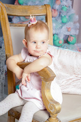 Fototapeta premium Portrait of cute little blond princess girl with a crown in beautiful dress sitting on vintage chair in studio decorated in Christmas theme with toys, tree and fireplace
