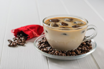 Glass bowl with ice coffee and red bag with crumbling coffee beans on white wooden table.