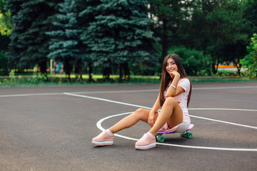 Fototapeta na wymiar Portrait of a smiling charming brunette female sitting on her skateboard on a basketball court.