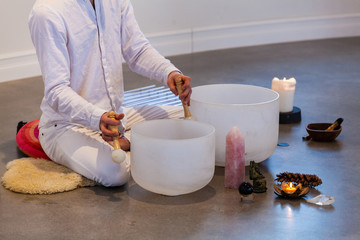 Man playing crystal bowls. Man dressed in white, sitting on a sheep skin with a close up on his hands playing crystal bowls with sacred objects displayed like stones, candles and more