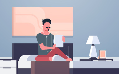 man listening audio book through headphones guy using tablet sitting on bed modern home bedroom interior male character full length flat horizontal