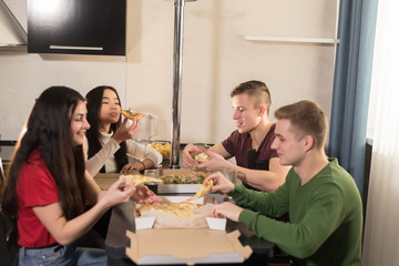 Company of four young friends sitting in kitchen and taking out pizza pieces