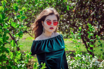 Portrait of beautiful young brunette woman with bright makeup, wearing emerald green velvet dress and bright red sunglasses, walking in a park