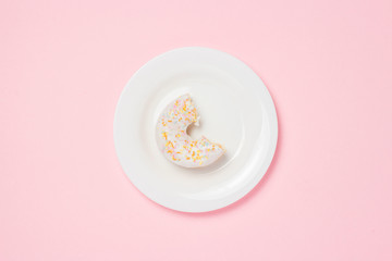 White plate with bitten off Fresh tasty sweet donut on a pink background. Bakery concept, fresh pastries, delicious breakfast, fast food, coffee shop. Flat lay, top view.