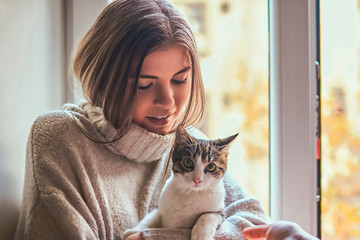 Beautiful girl in a warm sweater hugs her favorite cat sitting on the window sill next to the open window