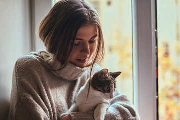 Beautiful girl in a warm sweater hugs her favorite cat sitting on the window sill next to the open window