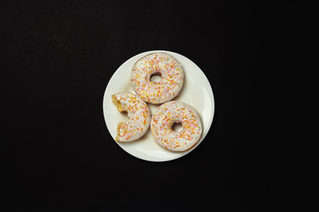 Delicious, sweet, fresh donuts on a white plate, black background. The concept of breakfast, fast food, coffee shop, bakery. Flat lay, top view.