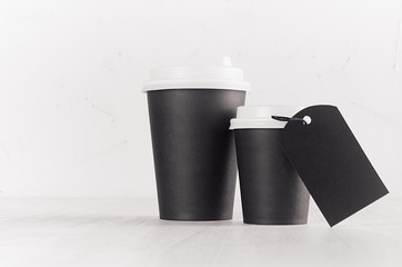 Coffee mockup - set of big and small black paper cups with white cap and blank label on white wood table with copy space, coffee shop interior. 