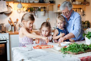 Kids and grandmother cooking italian pizza in cozy home kitchen for family dinner. Cute girls are preparing homemade food. Old senior woman is teaching three little sisters. Children chef concept.
