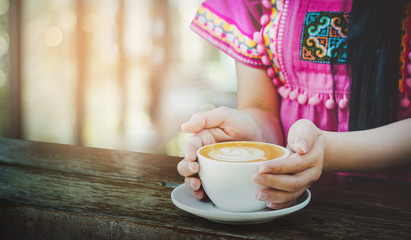 Asian woman wearing hmong dress holding cup of coffee on wooden table in cafe. Bokeh and blurred background with vintage light
