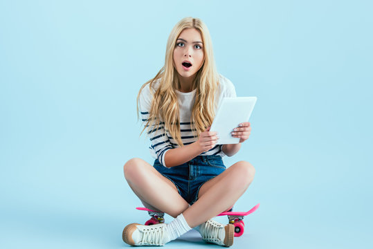 Shocked girl with digital tablet sitting on skateboard on blue background
