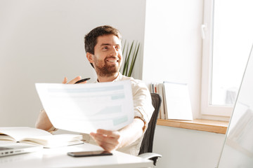 Image of businesslike office guy 30s wearing white shirt using laptop and paper documents, while working at modern workplace