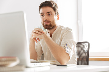 Image of european office guy 30s wearing white shirt using laptop and paper documents, while working at modern workplace