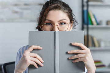woman in glasses covering face with book in modern office