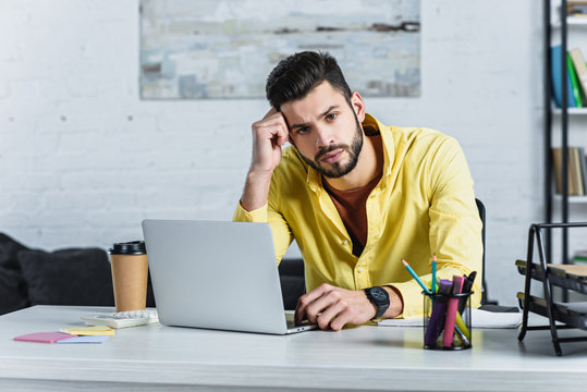 Thoughtful Bearded Businessman In Yellow Shirt Looking At Camera And Using Laptop