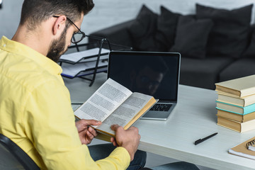 bearded man studying with book near laptop with blank screen in modern office