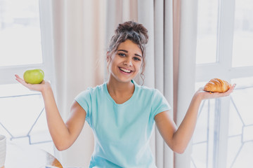 Beautiful young slender brunette with green apple in one hand and croissant in the other hand on background of vegetables on kitchen table making a difficult choice between healthy and harmful food. © Andrii