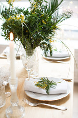 Beautiful springtime table setting with green leaves and mimosa branches, bright white table dinner decoration