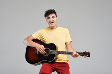 Handsome young man playing acoustic guitar and sings a fun song loudly. sitting on bar stool isolated on grey background