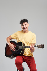 Handsome young man playing acoustic guitar and sings a fun song loudly. sitting on bar stool isolated on grey background