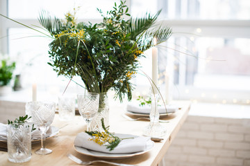 Beautiful springtime table setting with green leaves and mimosa branches, bright white table dinner decoration
