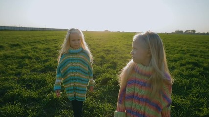 slow motion cute little blonde twin girls in striped oversize clothes stand on fresh green grass against evening sun