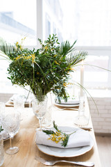 Beautiful springtime table setting with green leaves and mimosa branches, bright white table dinner decoration