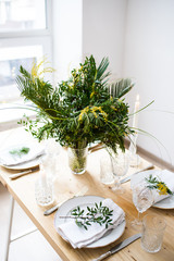 Beautiful springtime table setting with green leaves and mimosa branches, bright white table dinner decoration