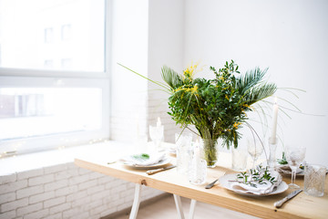 Beautiful springtime table setting with green leaves and mimosa branches, bright white table dinner decoration