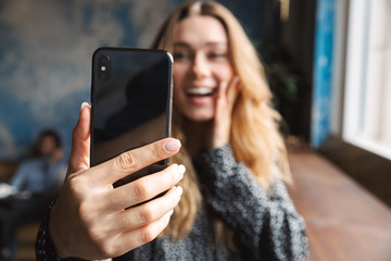Shocked excited beautiful young pretty woman sitting in cafe indoors take selfie by mobile phone.