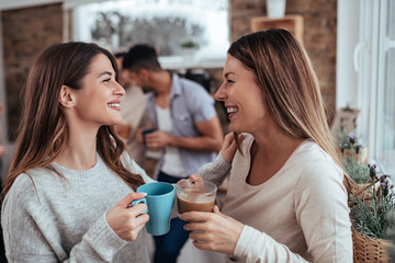 Two girlfiends in casual outfit talking and drinking tea and coffee. Smiling friends in modern cozy home having fun on winter day.