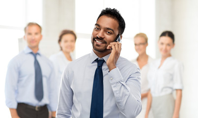 business, communication and people concept - smiling indian businessman calling on smartphone over colleagues on office background