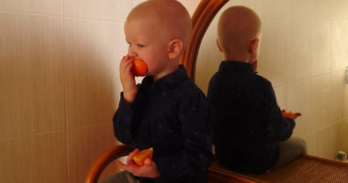Little blond boy sits on dresser with mirror and eats an orange. The child is having a good time at home.