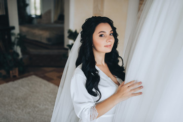 Beautiful bride is standing near the window in a white coat and smiling. Portrait of a cute brunette. Wedding photography.