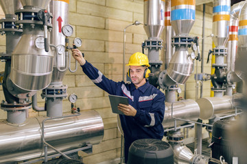 Young Caucasian worker in protective suit tightening the valve and using tablet while standing in...