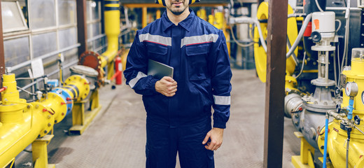 Young Caucasian worker in protective suit holding tablet and standing in heat plant.
