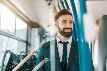 Close up of smiling bearded businessman in formal wear sitting in public transportation and looking through window.