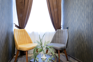 Beige and gray chairs near a wooden table with a vase and a houseplant on it,against a window with white tulle and coffee curtains.
