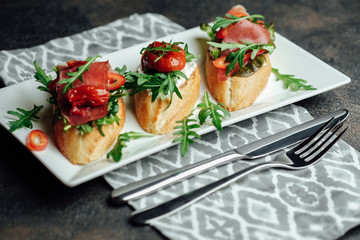 Italian bruschetta with prosciutto, dried tomatoes and arugula on a dark background.