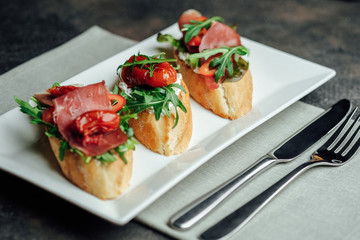 Italian bruschetta with prosciutto, dried tomatoes and arugula on a dark background.