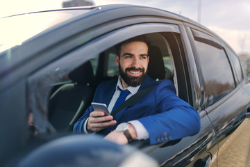 Close up of smiling businessman using smart phone while sitting in his car and leaning on the window.