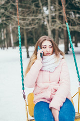 Young brown-haired woman alone on a swing in a park in the winter in the snowfall talking on a mobile phone - sadness and loneliness