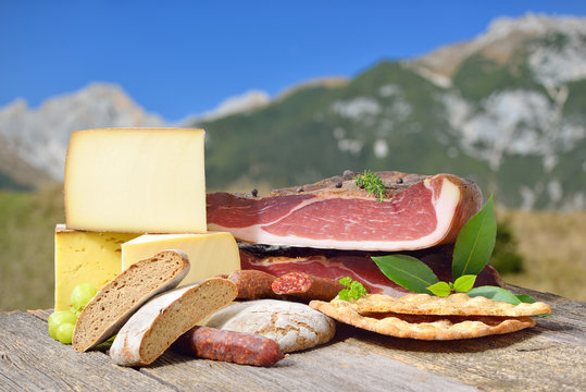 South Tyrolean specialties like bacon, sausages and cheese lying on a rustic table in front of mountains of the alps