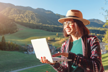 Fototapeta premium Happy successful young woman freelancer working remotely using a laptop in country cottage house with Alps mountains at the background
