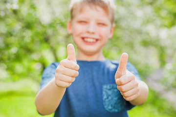 Handsome cheerful 9 years old boy giving two hands with thumbs up into camera as symbol of success. Closeup of fingers with focus at them and blurry face of blonde cute happy kid with great smile.