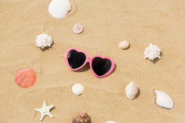 Fototapeta na wymiar vacation, valentine's day and summer holidays concept - pink heart-shaped sunglasses and shells on beach sand