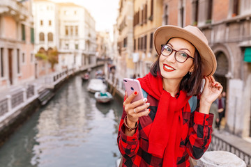 Fototapeta premium Young happy asian traveler woman with smartphone on the pier of a narrow canal and street in Venice. Vacation in Italy concept
