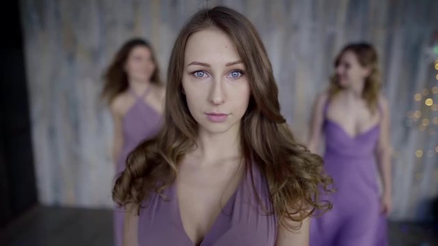 A beautiful girl stands in the middle, the other two girls go in the background and laugh. Three girls in delicate lilac dresses. Portrait close-up.