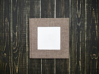 Closeup top view of family photo album with white frame in middle for text or image. Book isolated on brown wooden backgroumd. Horizontal flatlay photography. 