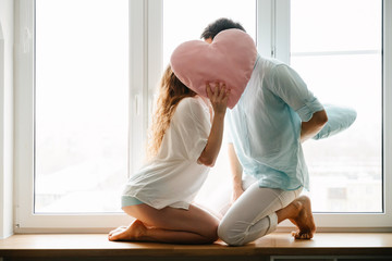 Couple girl and guy play with pillows near window.